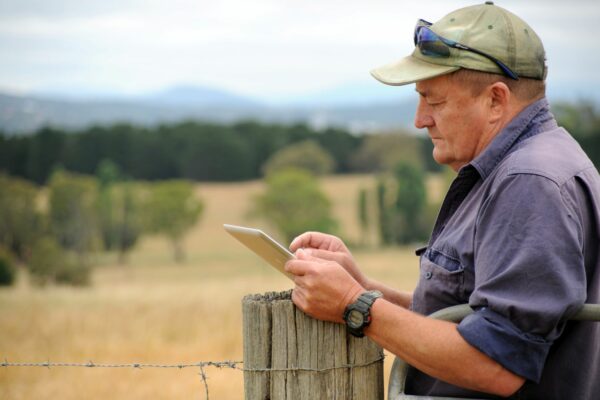 A man wearing a cap standing outside on a farm, leaning on a post using an ipad.
