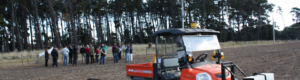 A field of dirt with threes in the background. At the front of the field is a red motorised cart. At the back of the field is a group of people.